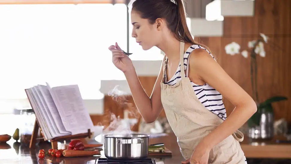 Person in an apron holding a spoon while reading a cookbook in the kitchen.