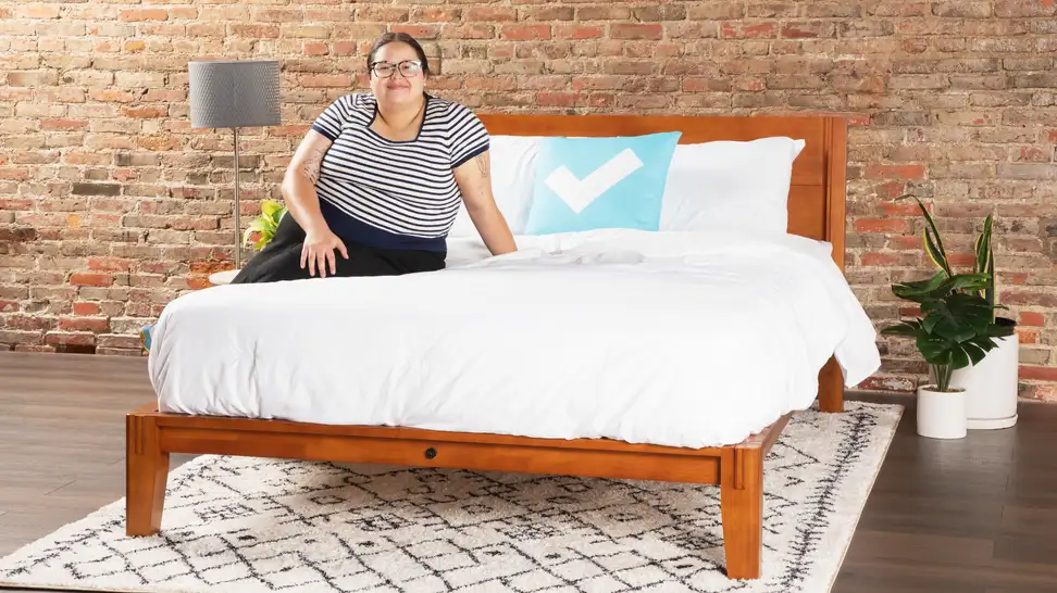 A woman sitting on a mattress atop The Bed by Thuma, a solid wood bed frame, against a brick wall.