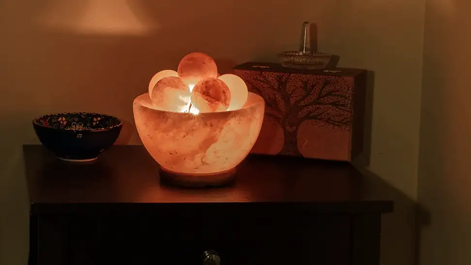A glowing orange Spantik Himalayan Fire Bowl Salt Lamp glows on a wooden table. It has a number of crystalline massage balls inside.