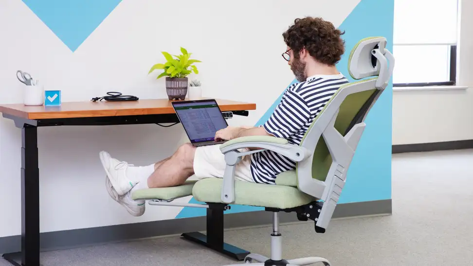 Person working with a laptop on their lab while slightly reclined in the green Edx Reclining Office Chair with Footrest sitting in front of an office desk.