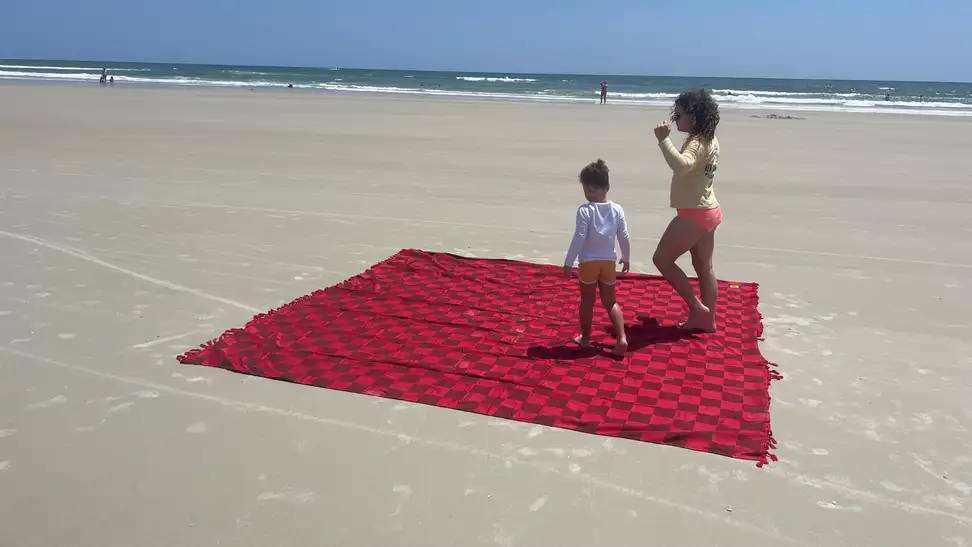 Two small children playing on top red checkered Big Blanket Big Beachy Blanket on beach shore.
