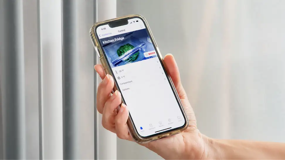 Person holding up smart phone to stainless steel refrigerator door with the Home Connect app on screen.