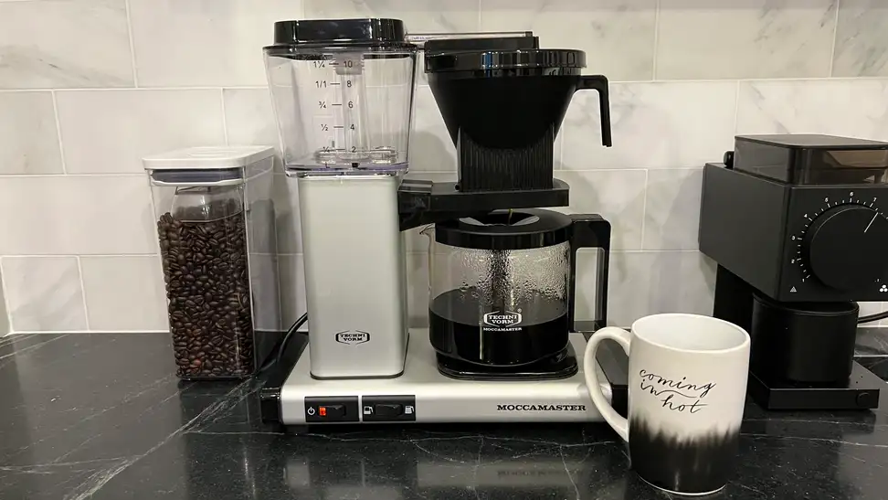 The Technivorm Moccamaster coffee pot full of coffee sitting on a black counter with a container of beans to the left, a coffee grinder to the right, and a mug in front that says "Coming in hot."