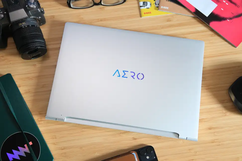 The Gigabyte Aero 14 OLED in silver, on a desk next to a camera and office supplies.