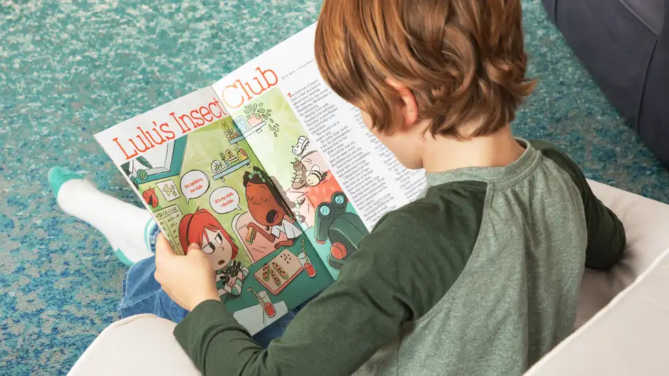 Child sitting in a small chair reading Highlights magazine.