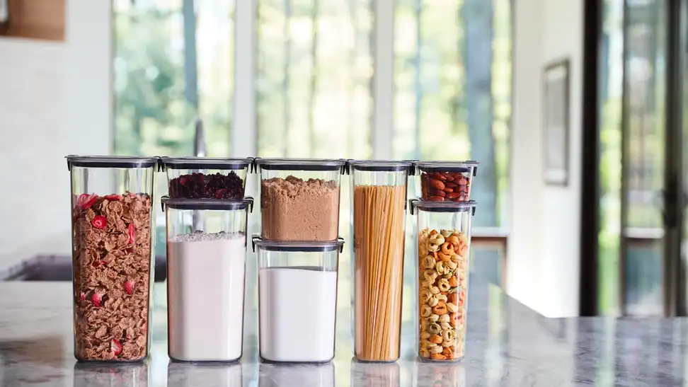On a countertop, several Rubbermaid Brilliance kitchen organizer bins are filled with dry cereal, tortellini, spaghetti noodles, sugar, spices, and so on.
