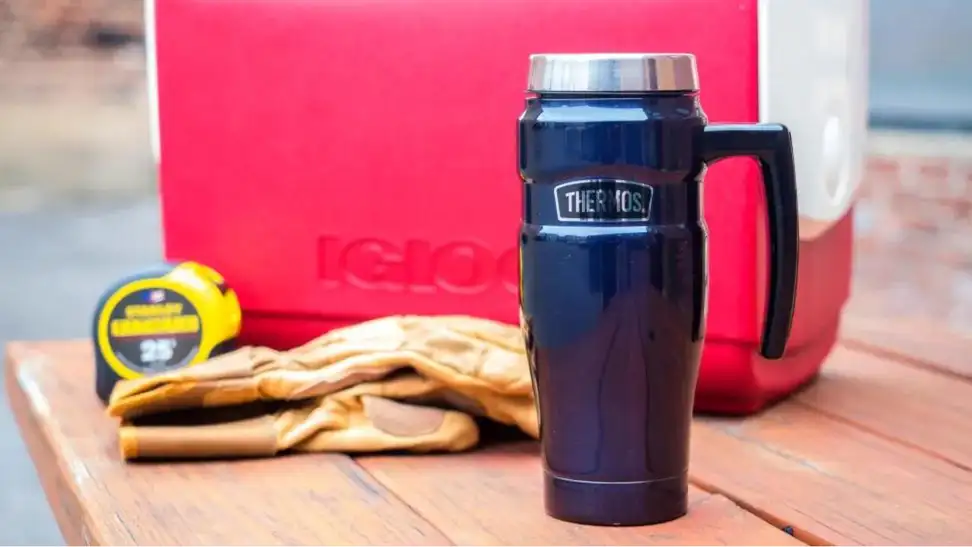 A Thermos travel mug sitting on a table next to a cooler.