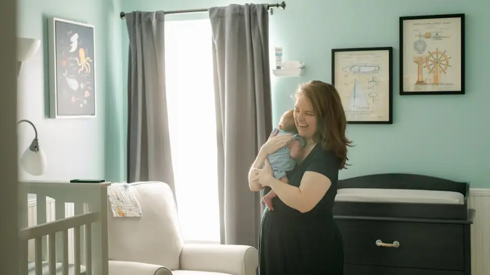Person smiling while holding newborn baby in arms inside of nursery room.