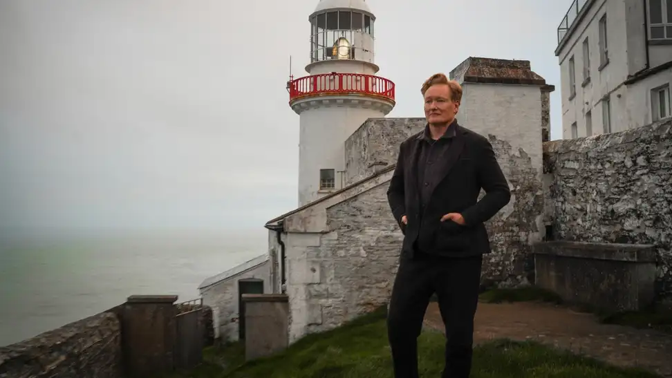 An image of Conan O'Brien standing on a shoreline alongside a lighthouse.