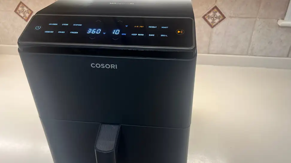The Cosori Dual Blaze Smart air fryer on countertop in front of tiled wall.