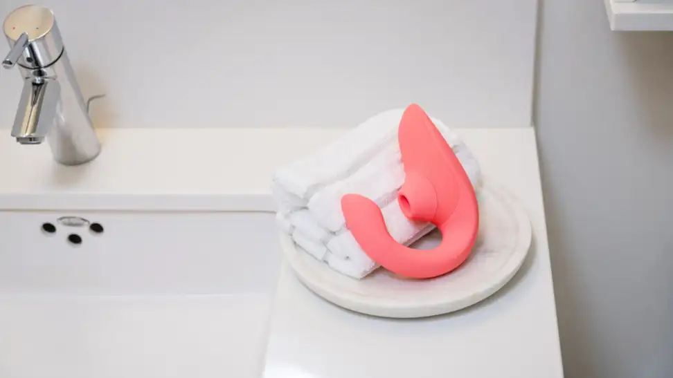 A Womanizer Blend in pink resting on hand towels on the side of a white bathtub.