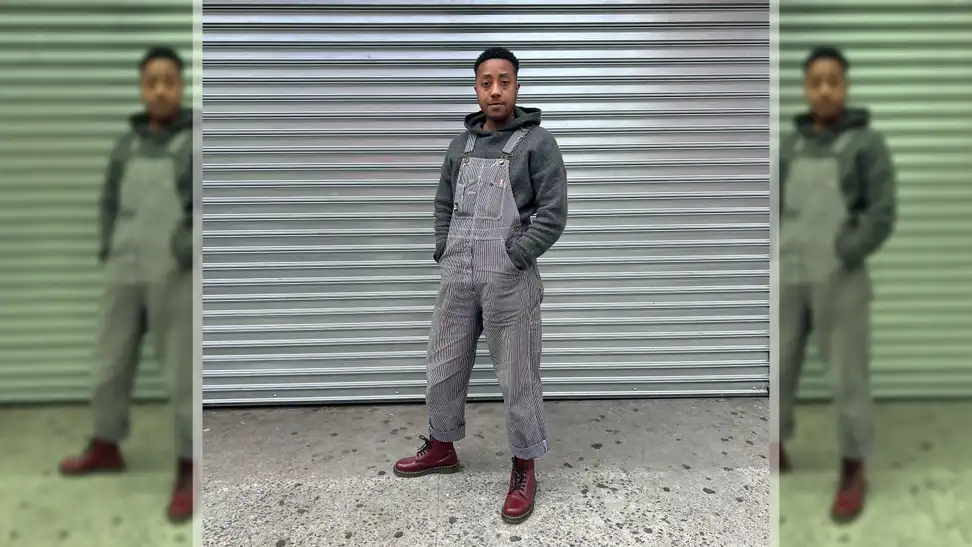 A person wearing overalls, red boots, and a gray hoodie, standing in front of a metal background.