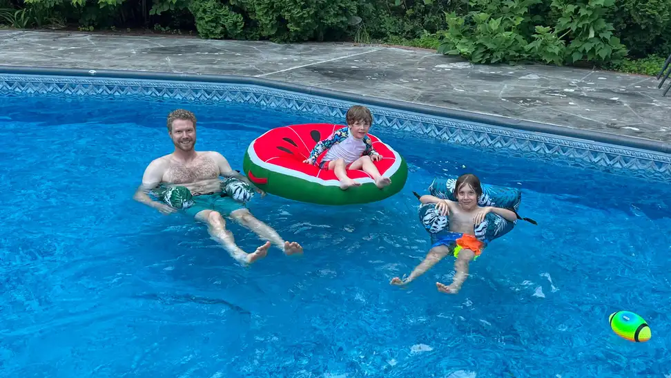 A man and child floating in pool chairs and a child floating on a Big Joe watermelon float in a pool.