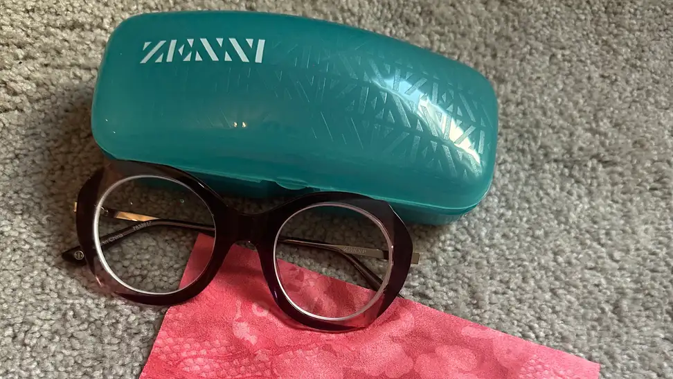 A pair of Zenni glasses next to a blue Zenni glasses case and pink fiber cloth.