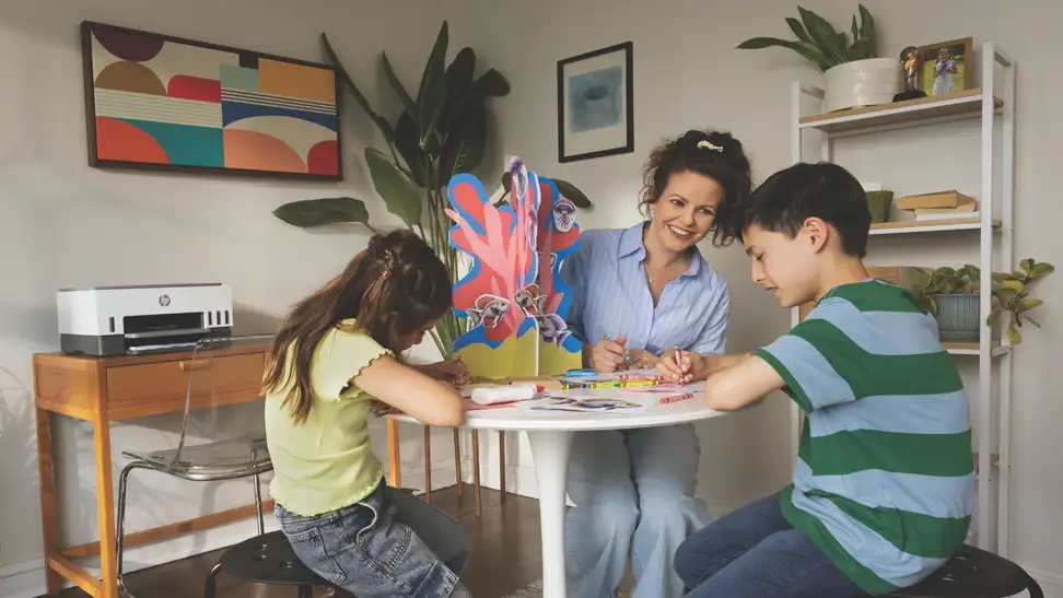 A woman and two children smile and work on a colorful arts and crafts project at a round table in a cozy, plant-filled home office with an HP printer in the background.