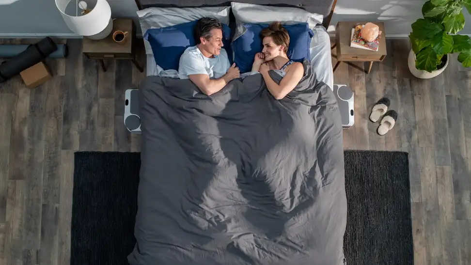 A couple lies in bed smiling at each other under a shared blanket, with Chilipad Dock Pro units visible on both sides of the bed.