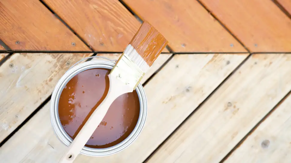 Deck boards, half of which have been stained. On the decking sits an open can of stain and a paintbrush.