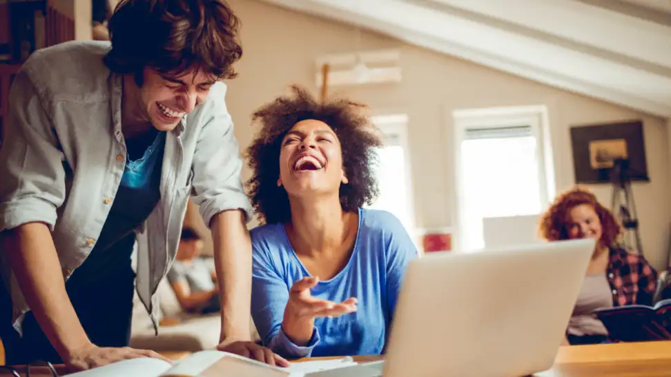 Two friends laughing together while working on a laptop at home, highlighting the fun and connectivity possible with T Mobile 5G Home Internet.