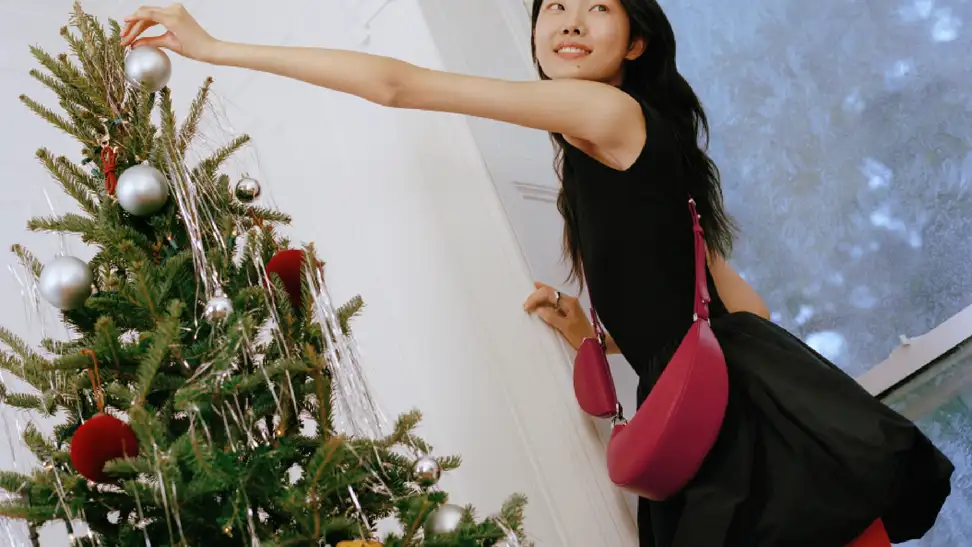Woman in black dress puts silver ornament on Christmas tree while wearing a red Kate Spade bag