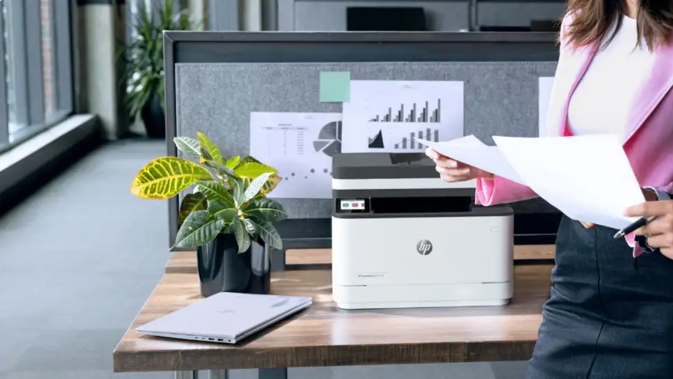 Woman in pink walks by a table with a printer, potted plant, and laptop