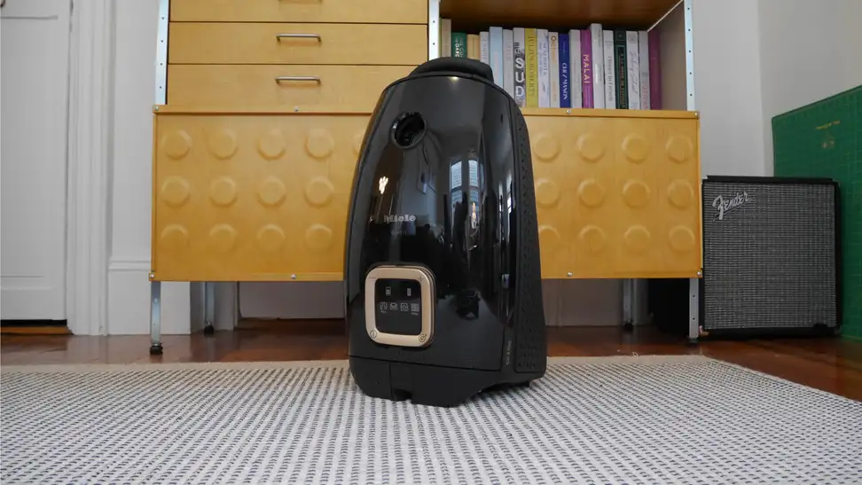 Black Miele Guard L1 Cat & Dog Vacuum sitting on a striped grey and white rug in front of tan drawers and a shelf.