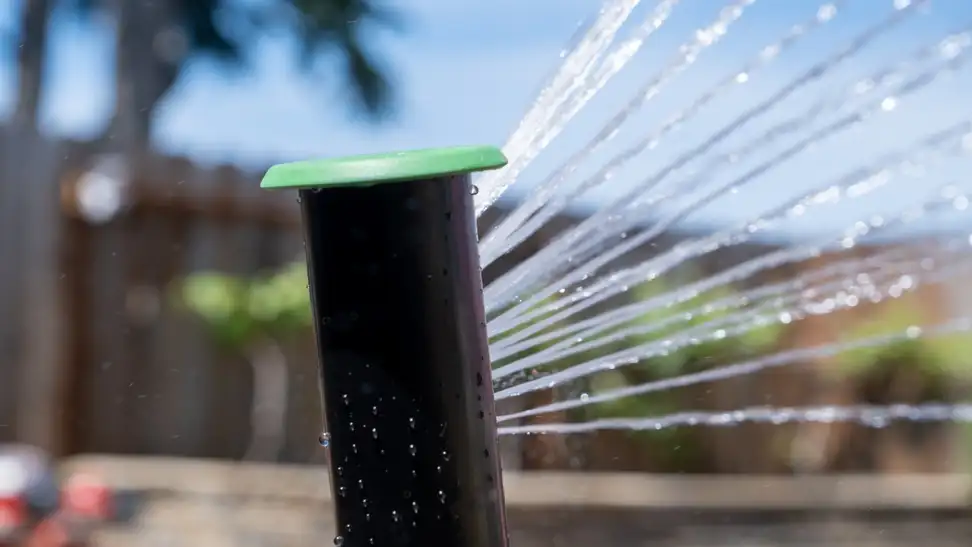 Irrigreen Smart Sprinkler 3 spraying water to the right, outside in front of a blue sky, tree, and building.