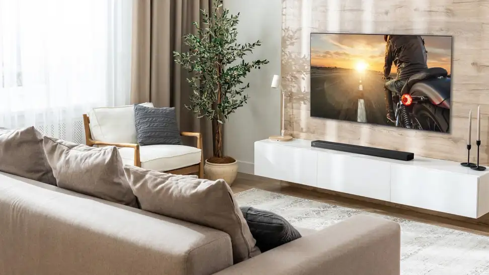 A bright, modern living room featuring an LG Dolby Atmos soundbar mounted beneath a large flat-screen TV on a light wood accent wall. The minimalist space includes a beige sectional sofa, white media console, indoor plant, and soft neutral decor, showcasing a clean and immersive home entertainment setup.
