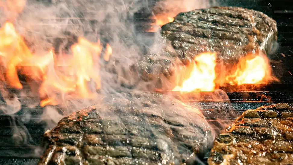 Steaks grilling over open flames with smoke rising.