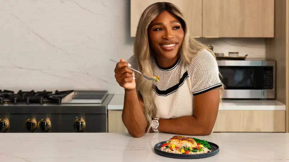 Serena Williams smiles while eating a Factor meal of blackened fish, grain salad, and green beans in a kitchen.