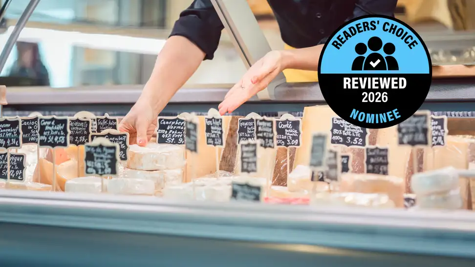 A deli case with an assortment of labeled cheeses, with a "Readers' Choice Reviewed 2026 Nominee" badge.