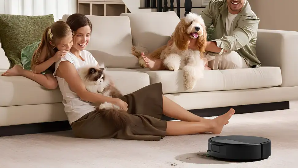 A woman and young girl sit on the floor with a Ragdoll cat while a man pets a fluffy dog on a cream sofa, as an iRobot Roomba robot vacuum cleans crumbs from the carpet beside them.