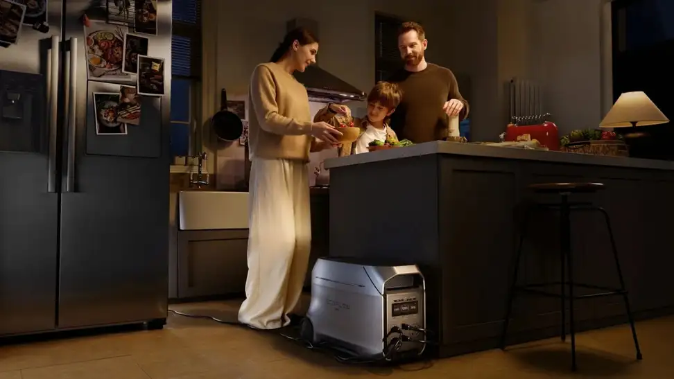 A family including a woman in a tan sweater and white pants stands next to a brunette male child and a man in a brown sweater, huddled around the kitchen counter. Their refrigerator, on the left side of the image and decorated with photos, is being powered by an EcoFlow device.