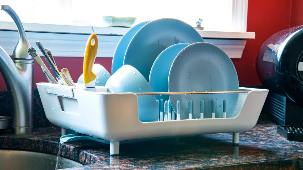 A dish rack filled with blue plates, cups and utensils sits next to a kitchen sink.