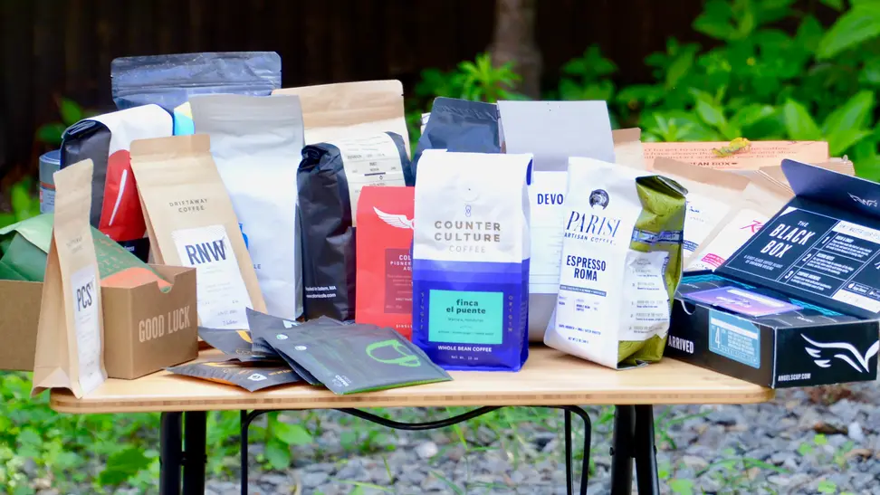 Over a dozen different bags of coffee beans arranged on a wood table outdoors.
