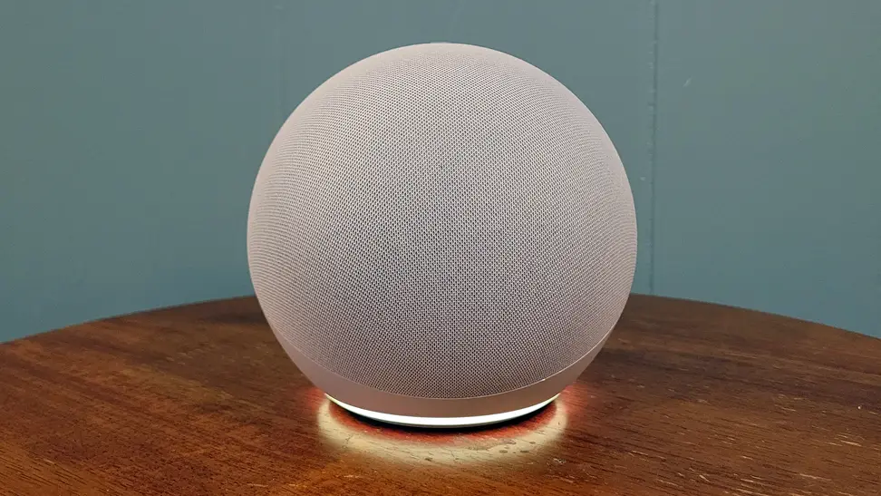 A round, gray smart speaker stands on a brown table agains a green wall
