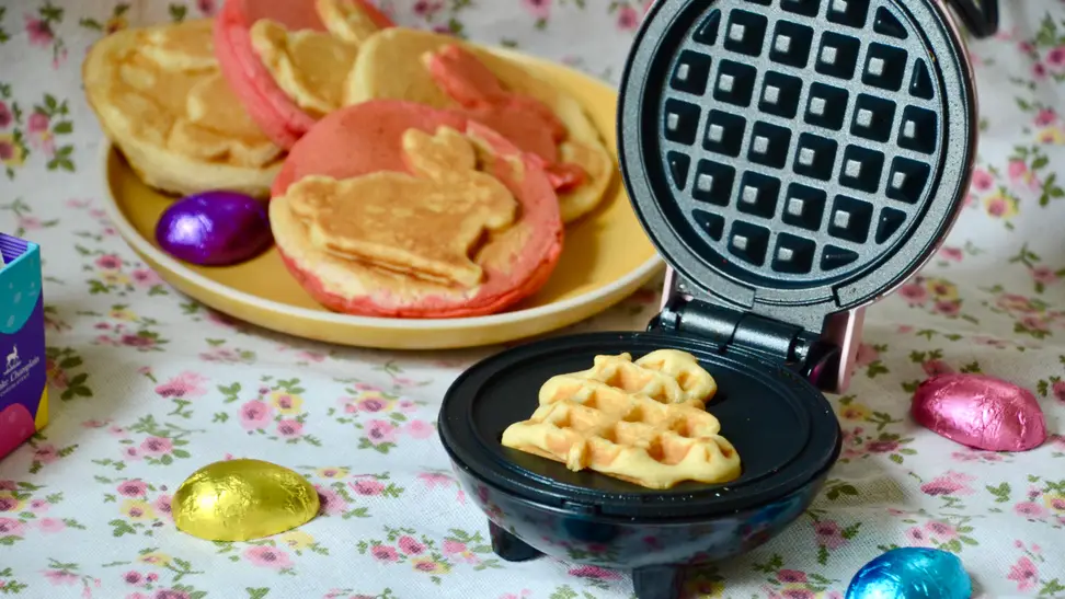 A bunny-shaped waffle is in the Dash Bunny mini waffle maker. Some cooked waffles are in the floral background, as are the Easter egg chocolate.