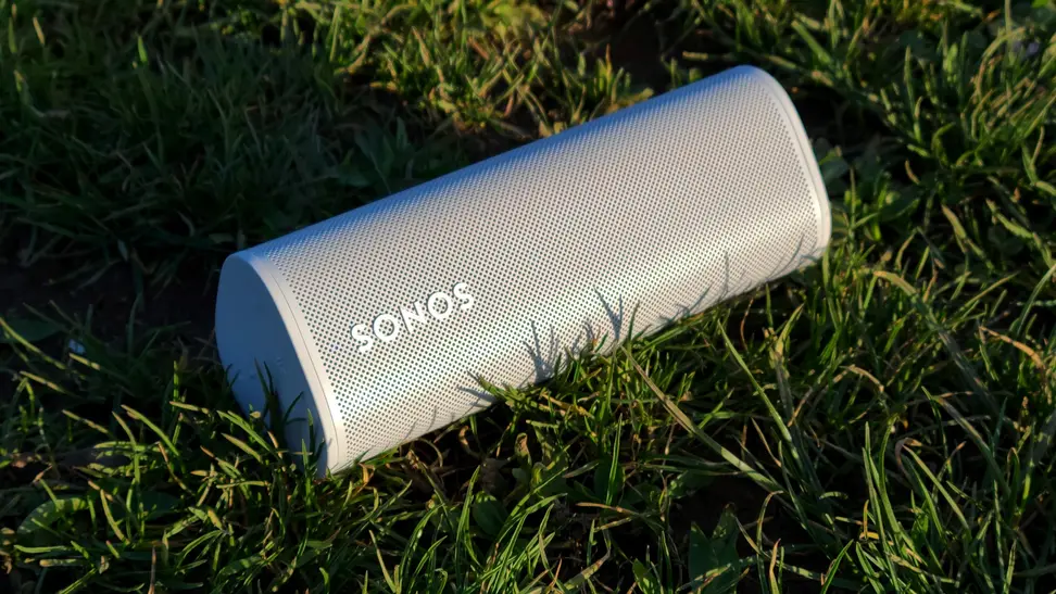 The tubular Sonos Roam speaker sits horizontally atop blades of grass as the sun sets
