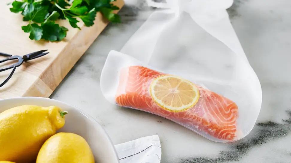 On a marble kitchen counter, a piece of salmon fillet and a slice of lemon are in an Anova silicone sous vide bag. Next to the bag, two lemons and some fresh herbs are scattered around.