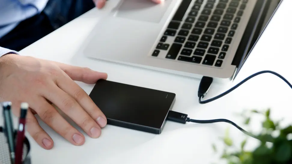 A hand reaches an external hard drive that's connected to a laptop on a desk.