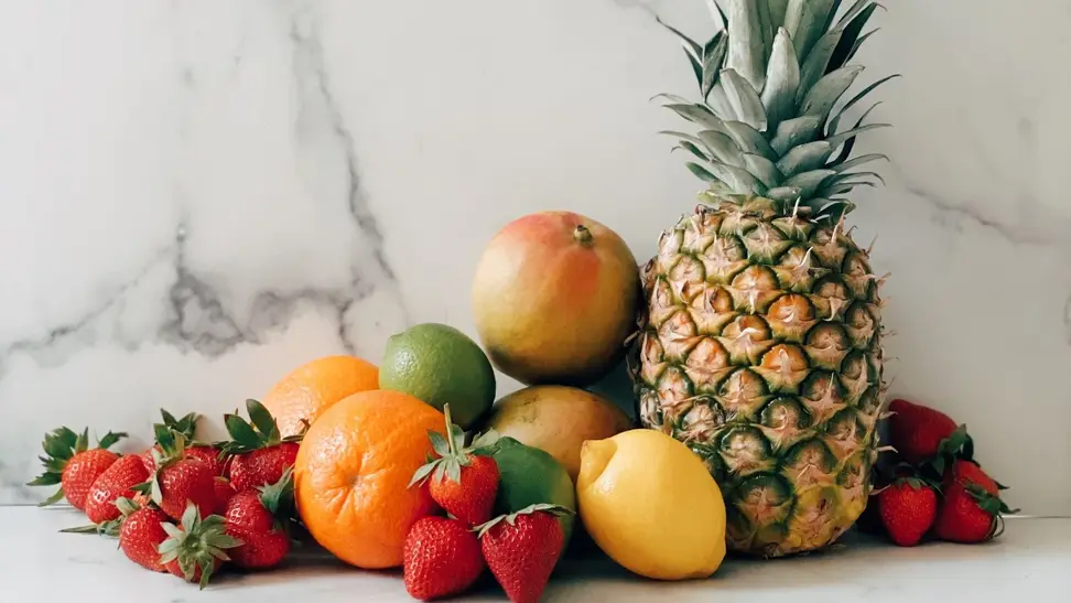 A pineapple, strawberries, mangoes, oranges, and lemons arranged in a pile on a white marble surface.