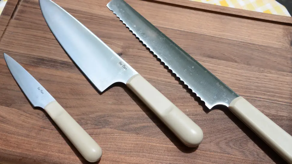 On a walnut wood cutting board by Our Place, a set of knives are on display. From the left, there's a paring knife, a chef's knife, and a serrated slicing knife.