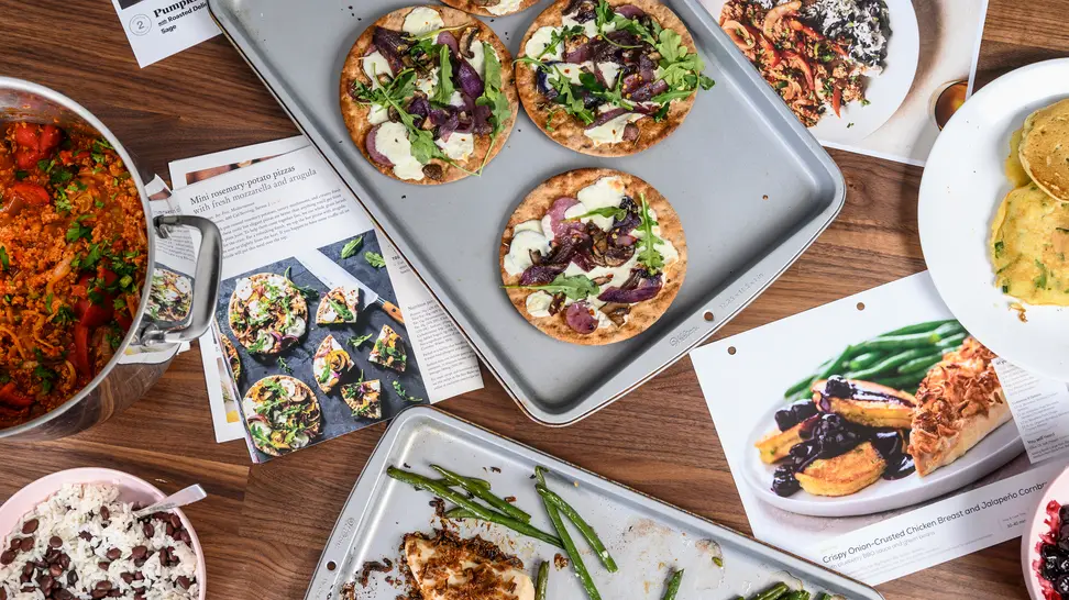 A selection of meal kit recipe cards and prepared food laid out on a table viewed from above.