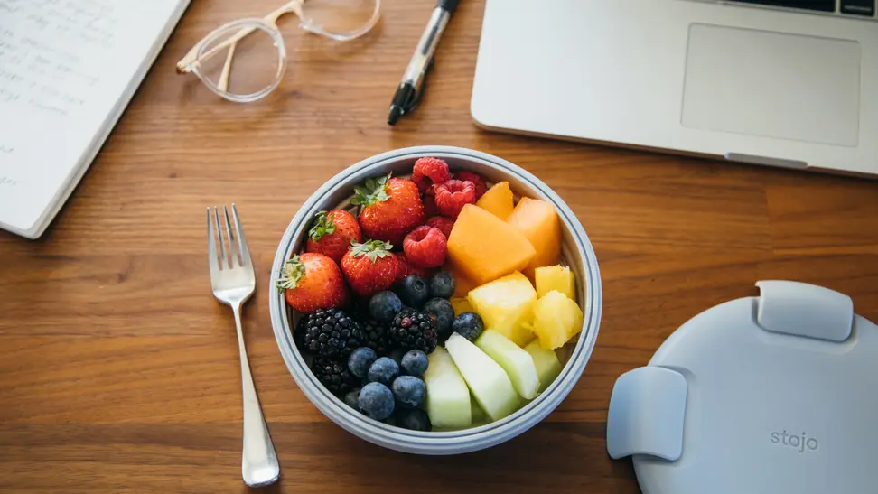 In the center of the desk, there's a Stojo bowl full of strawberries, blueberries, blackberries, cut orange, and other fruits. Beside the bowl, there's a fork. There's a notebook, a pair of glasses, a marker, and a MacBook scattered.