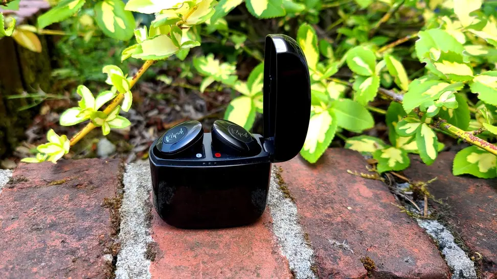 The black enameled Klipsch T5 II True Wireless ANC sit with their metallic case open on a brick ledge in front of greenery.