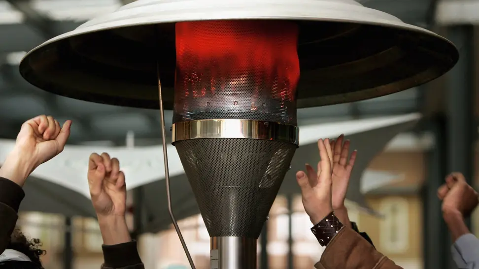 A small gathering of folks warm their hands around a glowing patio heater.