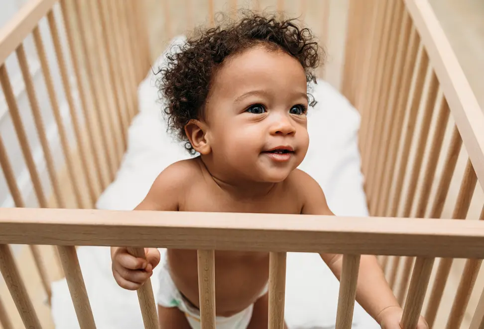 A small child stands up in a wooden crib.
