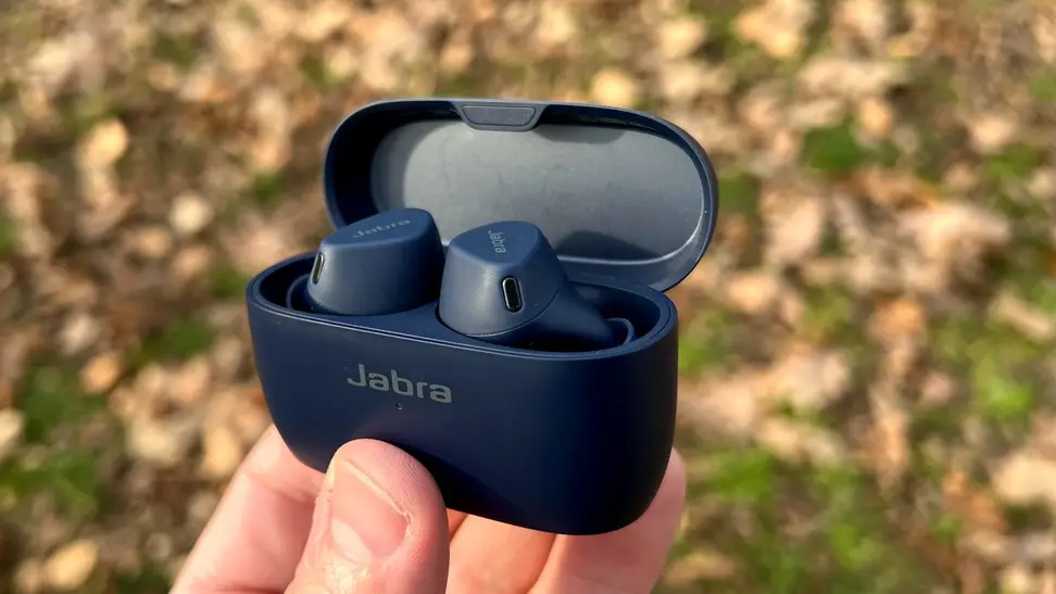 A pair navy earbuds sit in their open charging case, held in a hand before a leaf-covered ground.