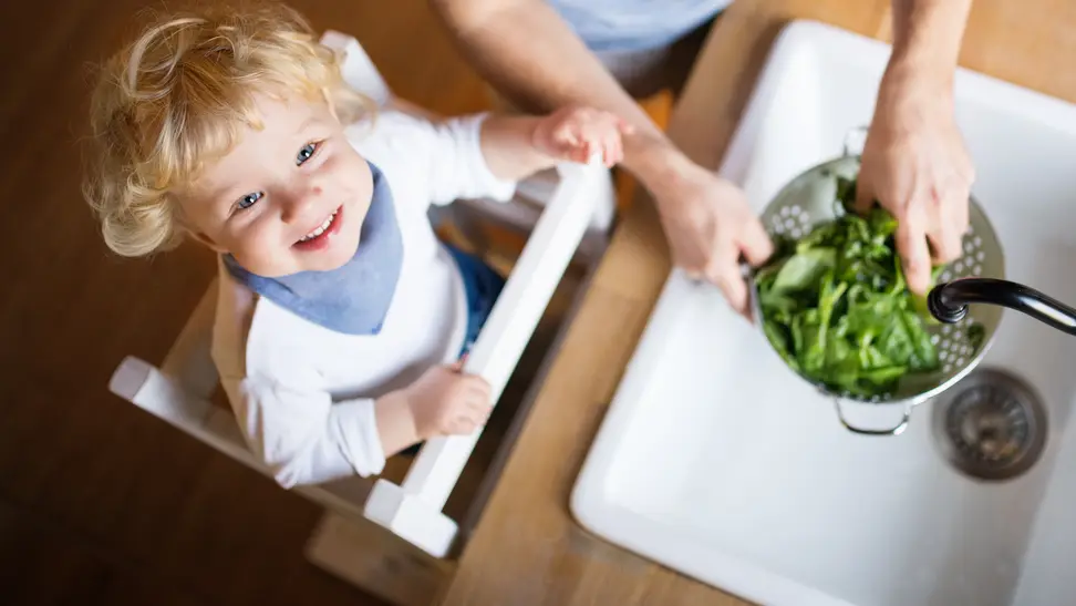 A small child standing in a learning tower at the kitchen sink helping a parent wash vegetables.