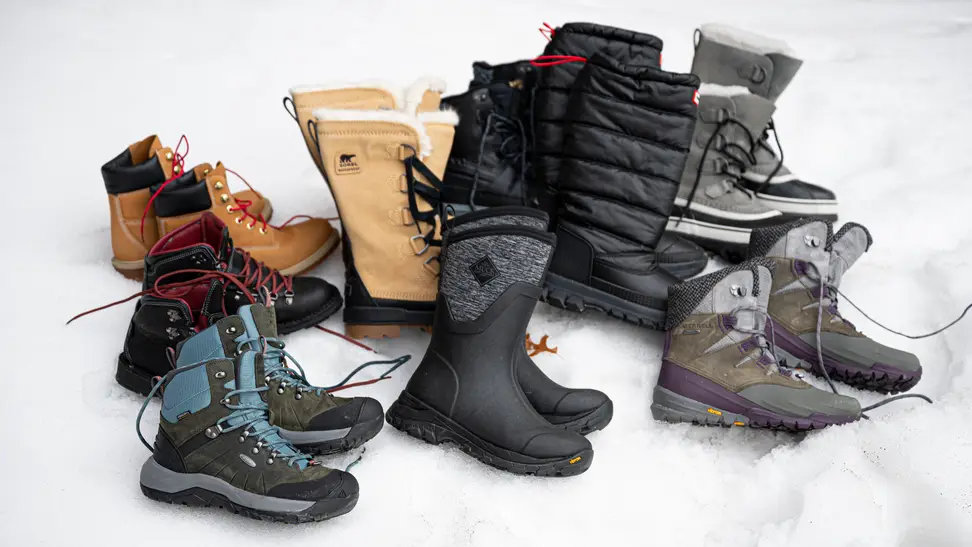 A number of pairs of winter boots, from various brands, laid out on a bed of snow.