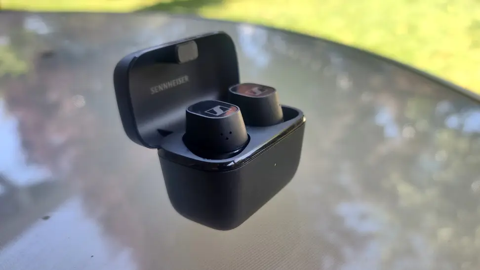 Sennheiser's all-black CX Plus headphones sit on a glass table on a grassy lawn in their case.